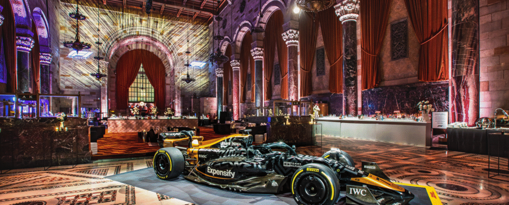 A luxurious event space with grand architectural features, including vaulted ceilings, arched windows, and ornate columns bathed in colorful lighting. At the center of the room, a sleek black and gold Formula 1 race car with prominent branding is displayed on a branded platform. The floor features intricate mosaic tile patterns, while the background includes elegant red drapery, modern cocktail bars, floral arrangements, and lounge areas. The dramatic lighting and mix of historic architecture with modern elements create a striking and sophisticated atmosphere.