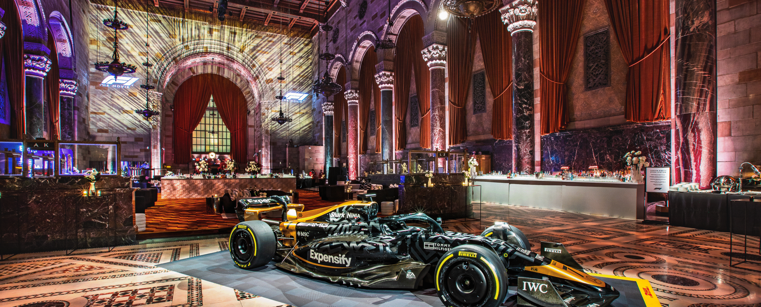 A luxurious event space with grand architectural features, including vaulted ceilings, arched windows, and ornate columns bathed in colorful lighting. At the center of the room, a sleek black and gold Formula 1 race car with prominent branding is displayed on a branded platform. The floor features intricate mosaic tile patterns, while the background includes elegant red drapery, modern cocktail bars, floral arrangements, and lounge areas. The dramatic lighting and mix of historic architecture with modern elements create a striking and sophisticated atmosphere.